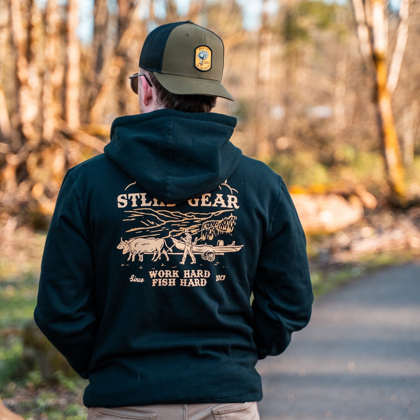 Person wearing a dark hoodie with STLHD Gear WORK HARD FISH HARD logo and text, standing in a forest.