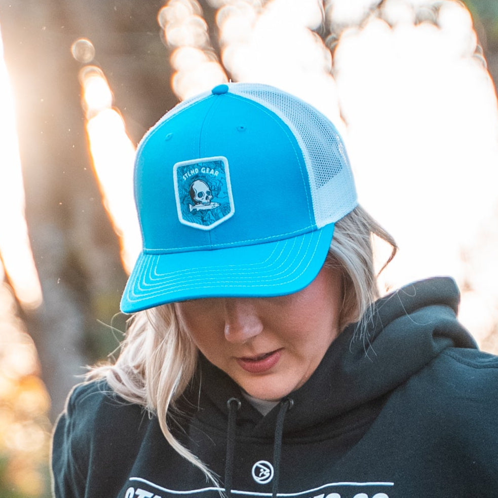 Person wearing a blue cap with a  STLHD GEAR skull and fish patch logo, blurred background