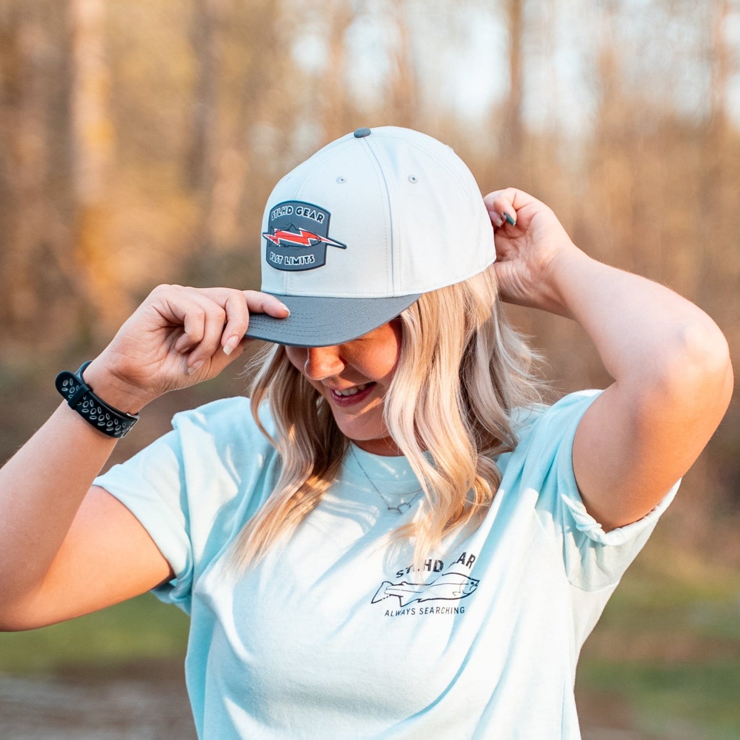 A blonde woman smiles as she adjusts her STLHD Fast Limits Hat by STLHD GEAR with one hand. She wears a light blue T-shirt and stands outdoors, trees softly blurred in the background.