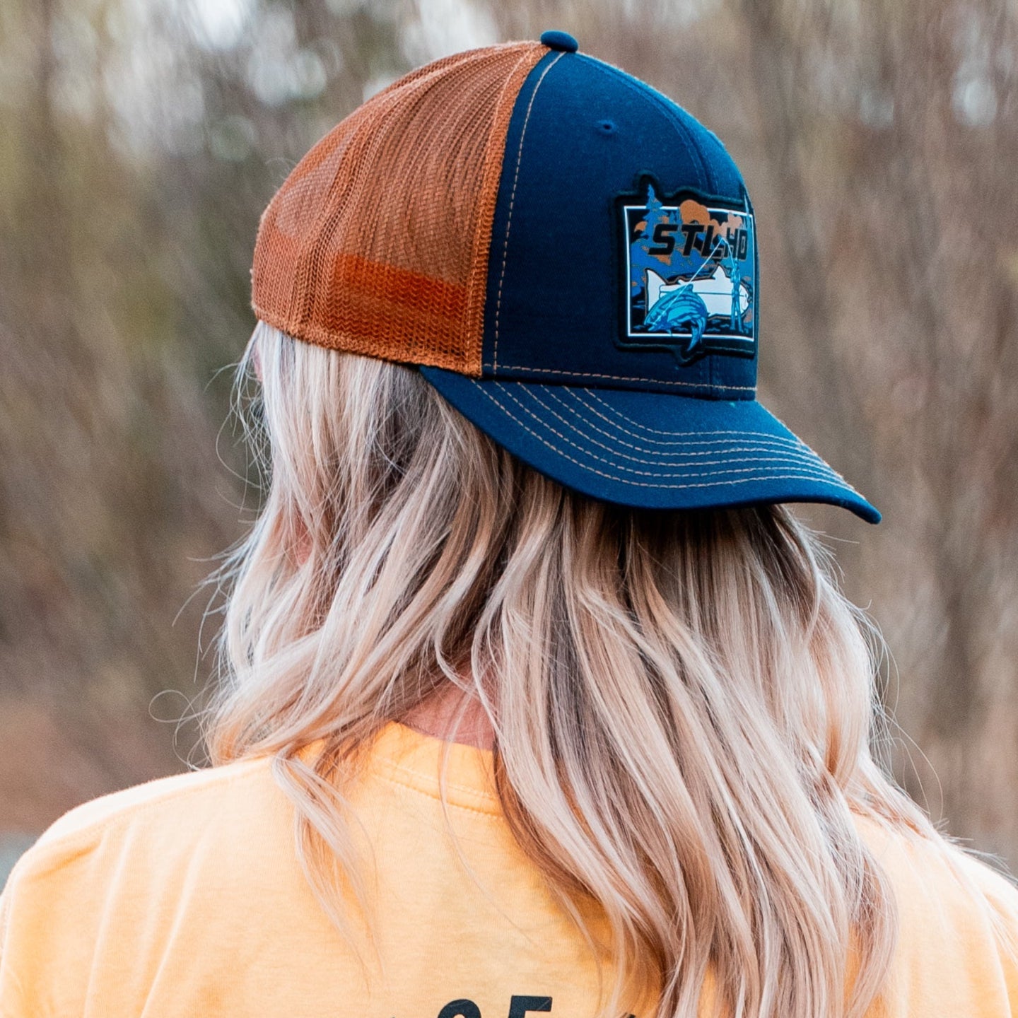 A person with long, wavy blonde hair, wearing a yellow shirt, faces away outdoors. They wear the STLHD GEAR Let’er Rip Trucker Hat—blue and brown with an adjustable snapback, a fish patch, and text. The background is blurred.