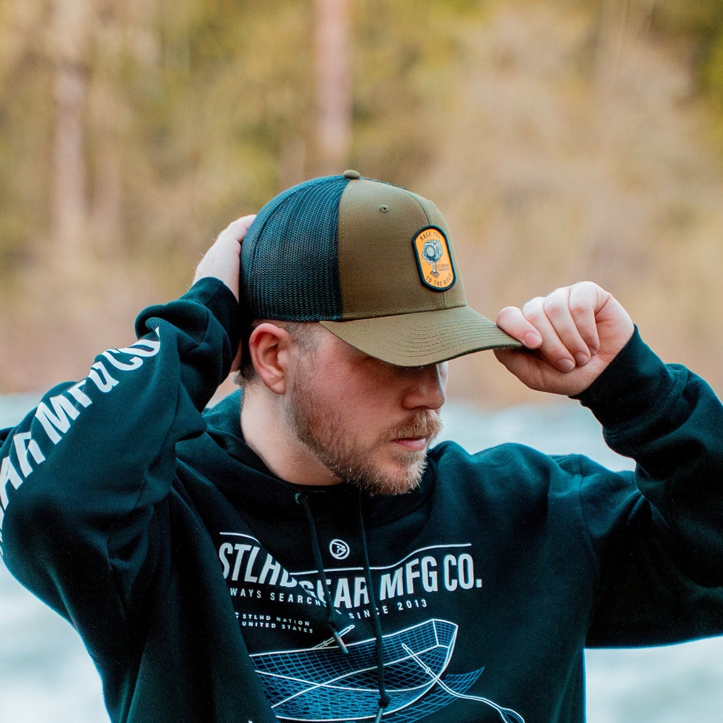 A man in a black hoodie and an STLHD GEAR Race Ya Flat Bill Trucker Hat with adjustable snapback stands outdoors, one hand on his hat, the other behind his head. Blurred trees and water form the background.