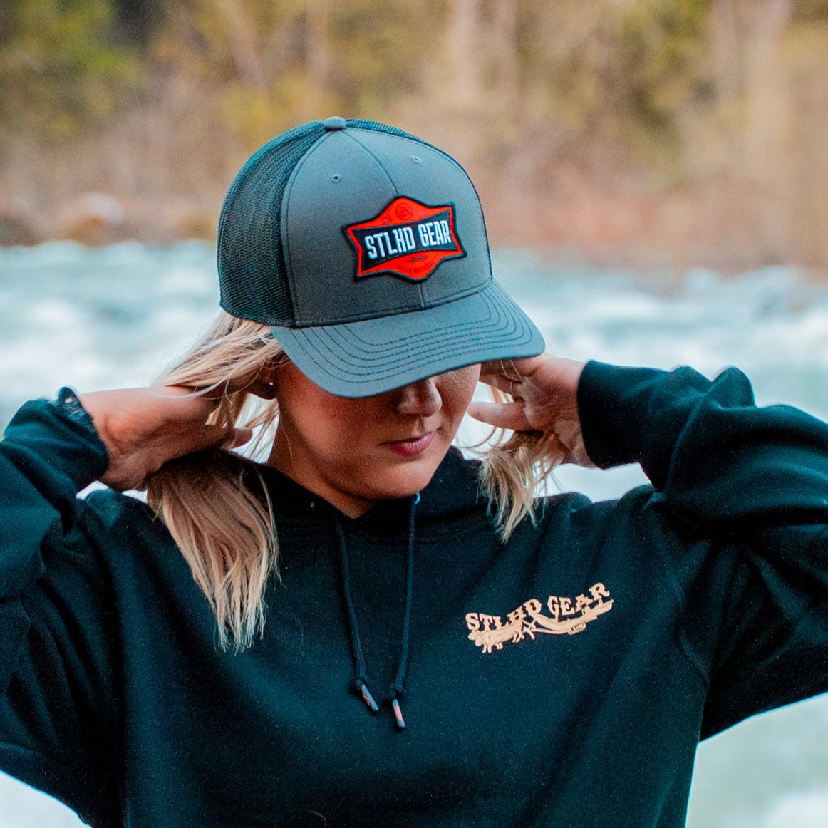 A person by a river adjusts the STLHD GEAR Never Not Fishing Trucker Hat, featuring the brand’s logo. They wear a dark hoodie as blurred trees and water fill the background.
