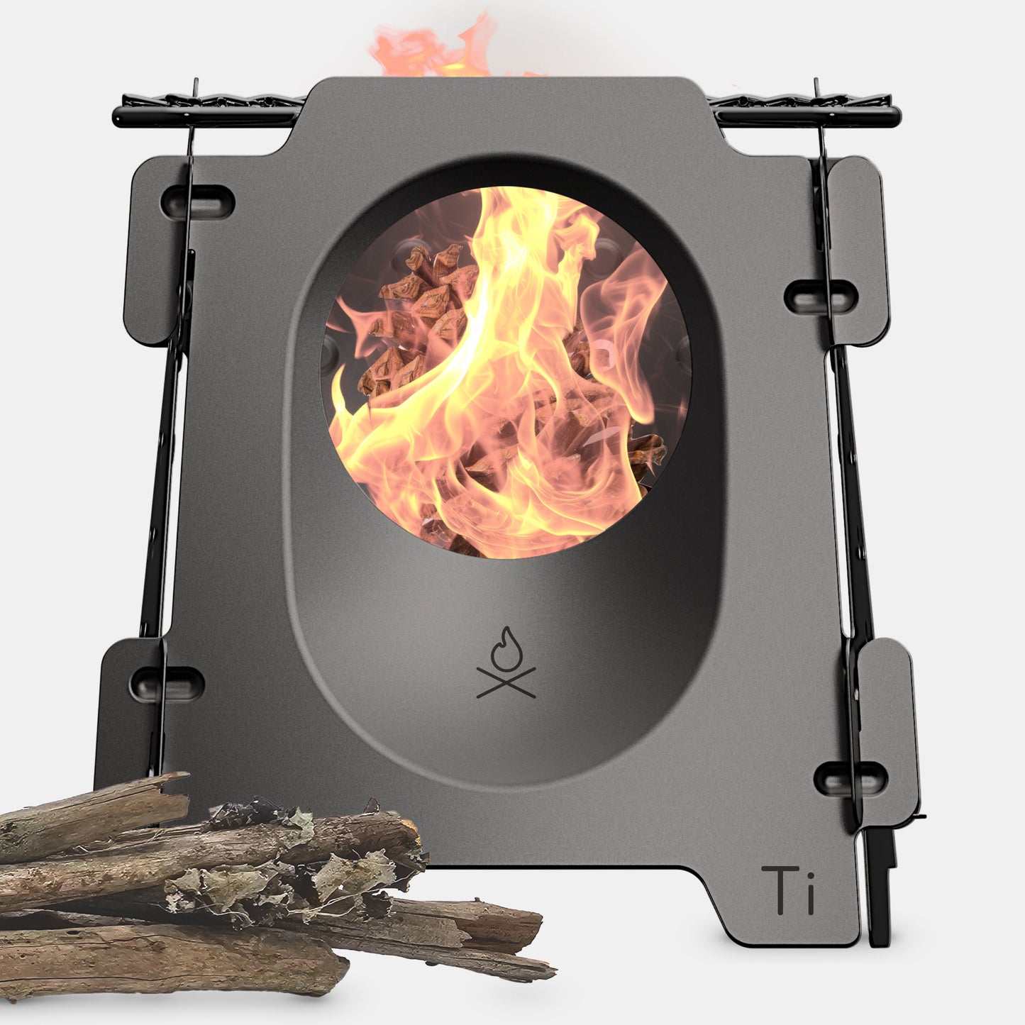 The Überleben Stöker Flatpack Stove by Überleben is a compact, rectangular metal wood-burning stove with a central oval opening displaying a bright fire. A small bundle of sticks sits beside this survival-grade tool, which features Ti labeling in the corner.