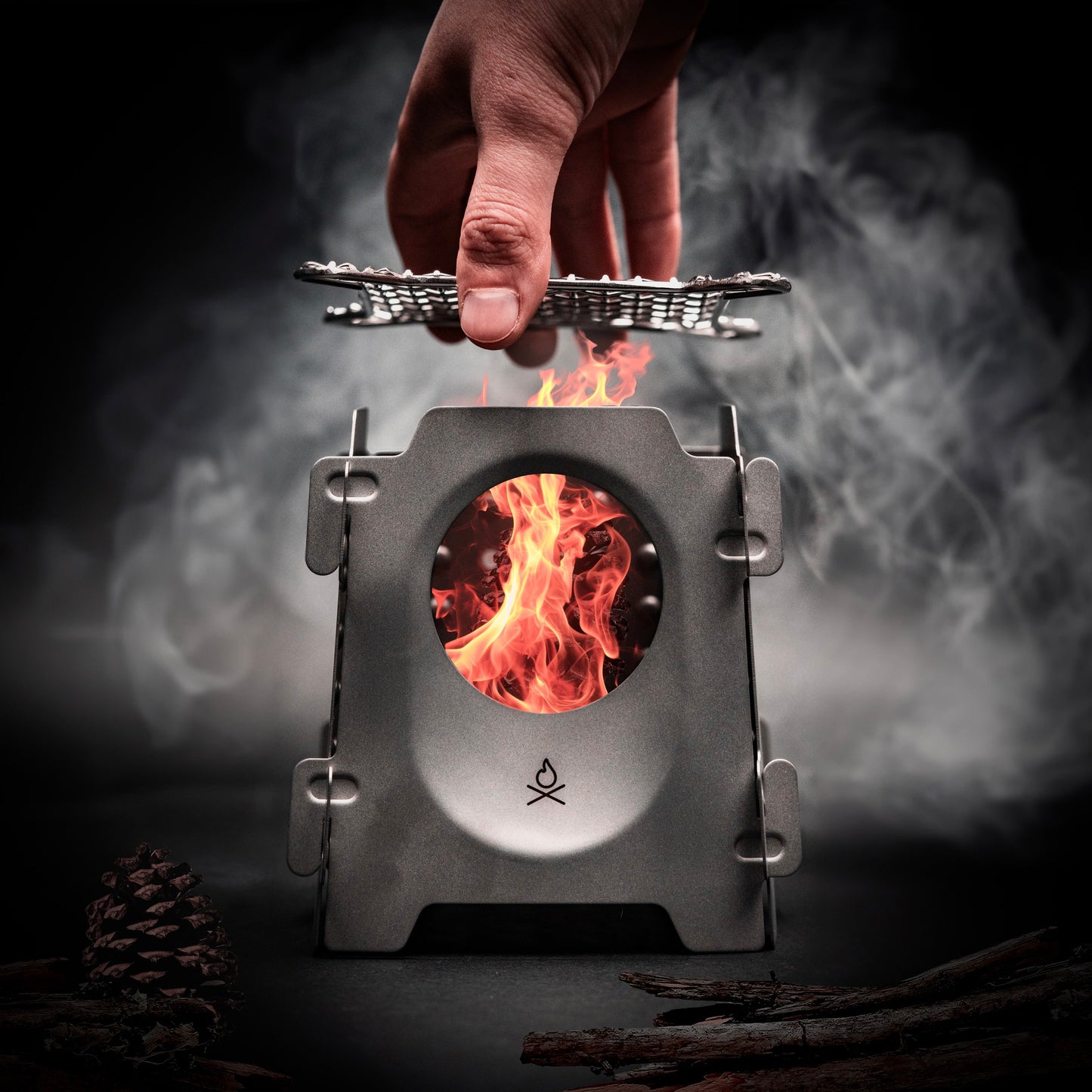 A hand sets a metal grate on the Überleben Stöker Flatpack Stove by Überleben, as flames glow inside. Smoke wafts up with pine cones and twigs nearby, all against a dark backdrop.