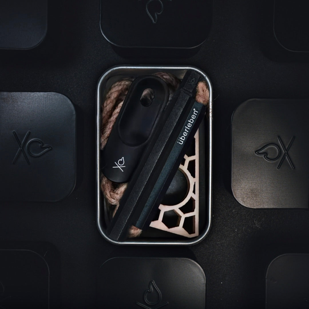 The Überleben FIREKIT – All-in-One Fire Starter Kit by Überleben comes in a compact metal tin and includes a black fire starter, rope, and an Überleben tool, displayed on a dark background with minimalist flame icons.