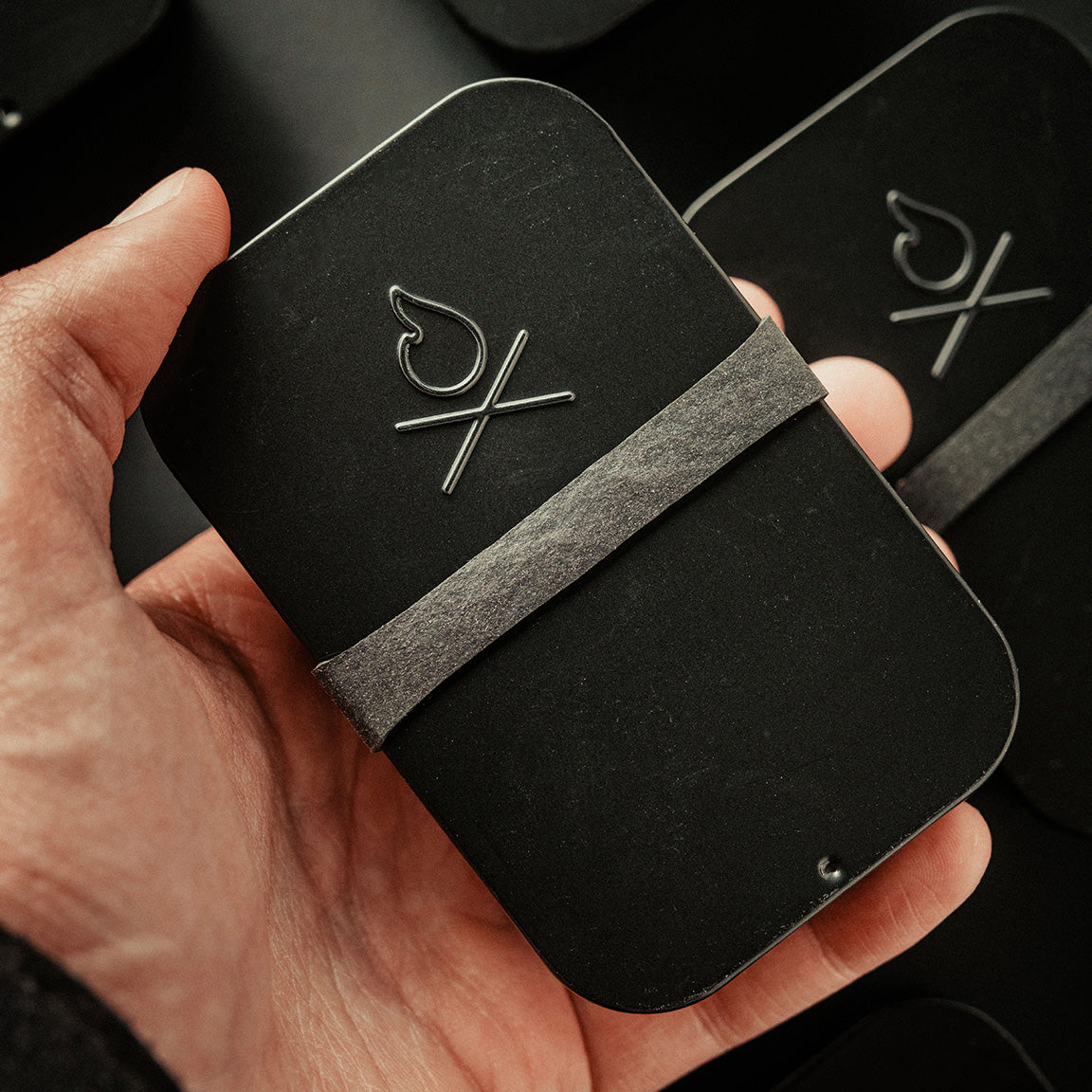 A hand holds the Überleben FIREKIT – All‑in‑One Fire Starter Kit, a sleek black tin with an embossed flame and X logo, secured by a gray paper band. Other Überleben tins sit in the background on a dark surface.