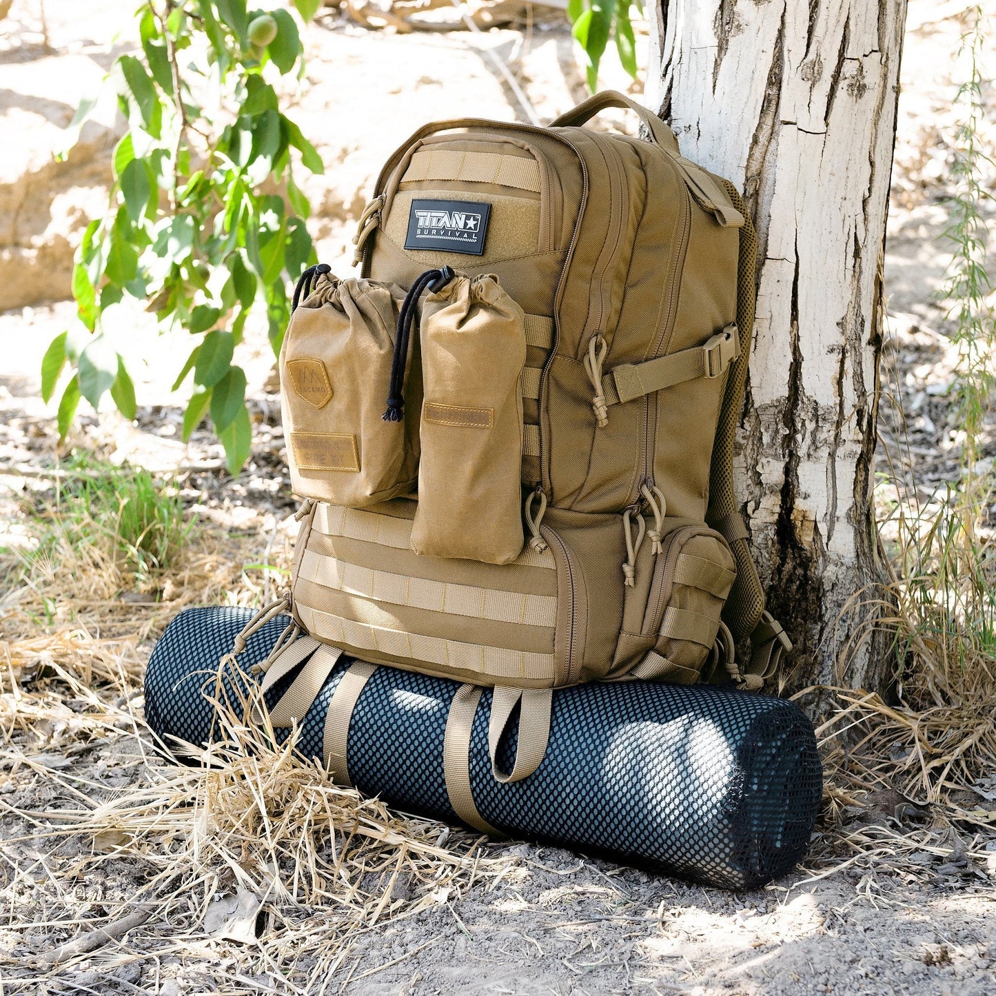 The TITAN Survival TACAMO BC50 72-Hour Tactical Backpack in tan, featuring two front pouches and a black sleeping mat attached underneath, rests against a white tree outdoors amid dry grass and green leaves.