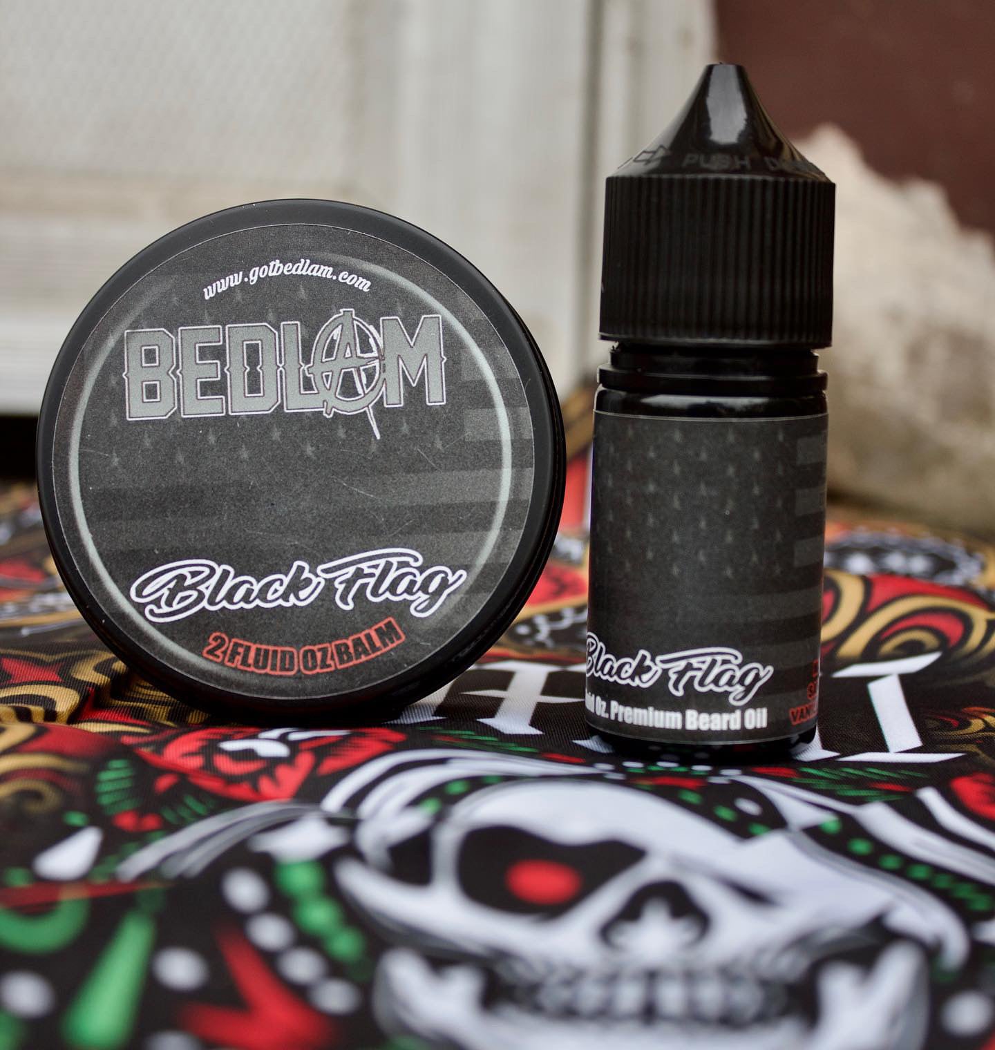 A tin of Bedlam Beard Companys BLACK FLAG Beard Balm and a bottle of BLACK FLAG Beard Oil rest on skull-patterned fabric, featuring black labels with white and red text and a bold, masculine scent.