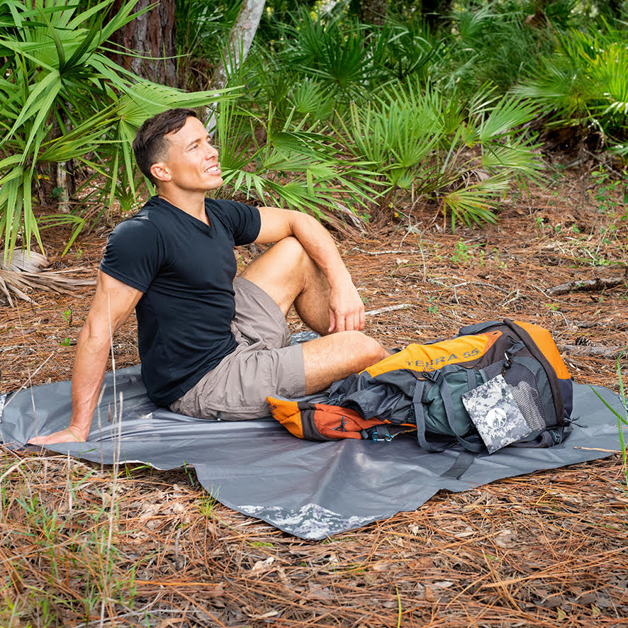 A man in a black t-shirt and khaki shorts smiles while sitting on the Pike Trail Pocket Blanket by Pike Trail in a forest clearing, with his hiking backpack beside him amid pine needles and green foliage.