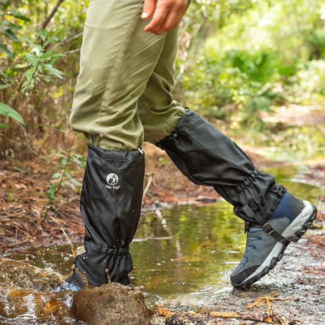 Wearing khaki pants, hiking shoes, and Pike Trail All-Season Waterproof Leg Gaiters—built for the trail, the hunt, and everything in between—a person walks through a shallow, muddy forest stream.