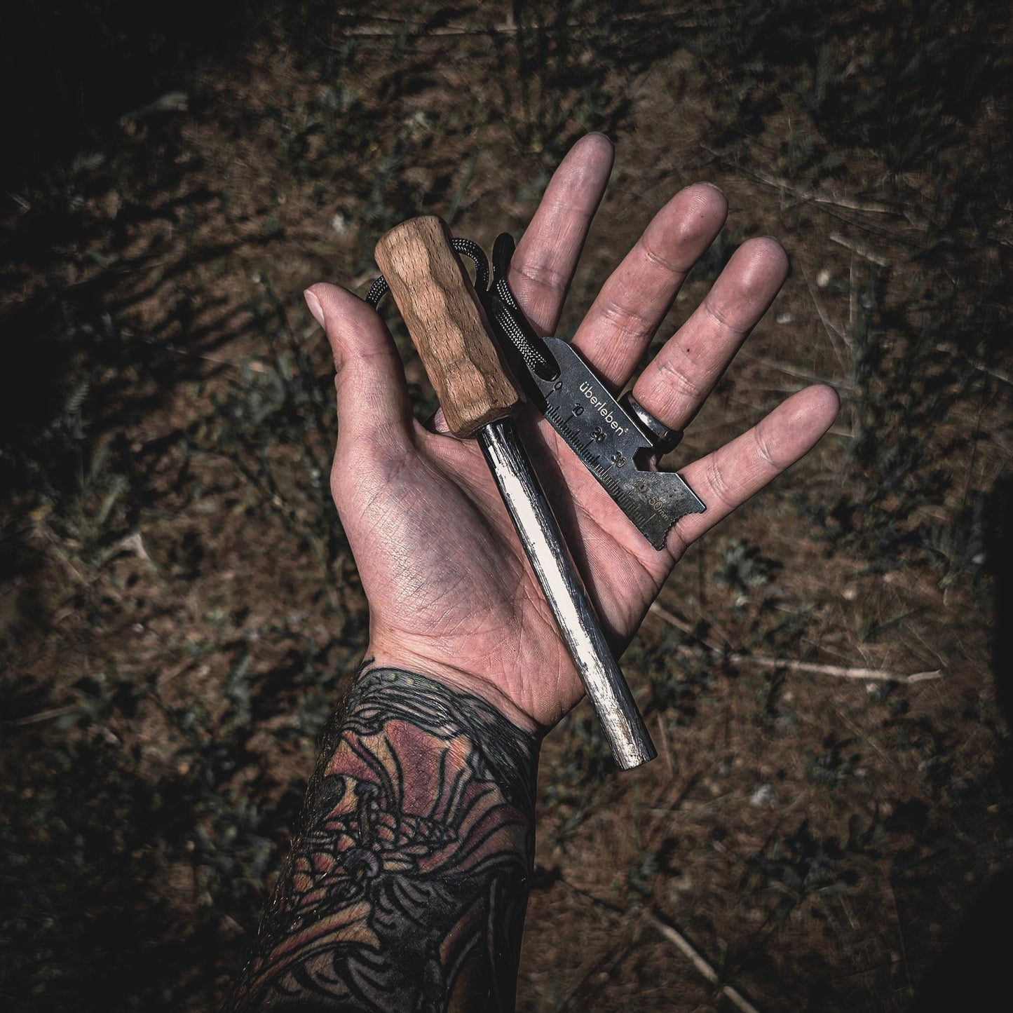 A tattooed hand holds the Überleben Kräftig Grip (PRO) – The Heavy-Duty Ferro Beast by Überleben, featuring a large ferrocerium rod with a wooden handle and black metal striker, outdoors over grass.