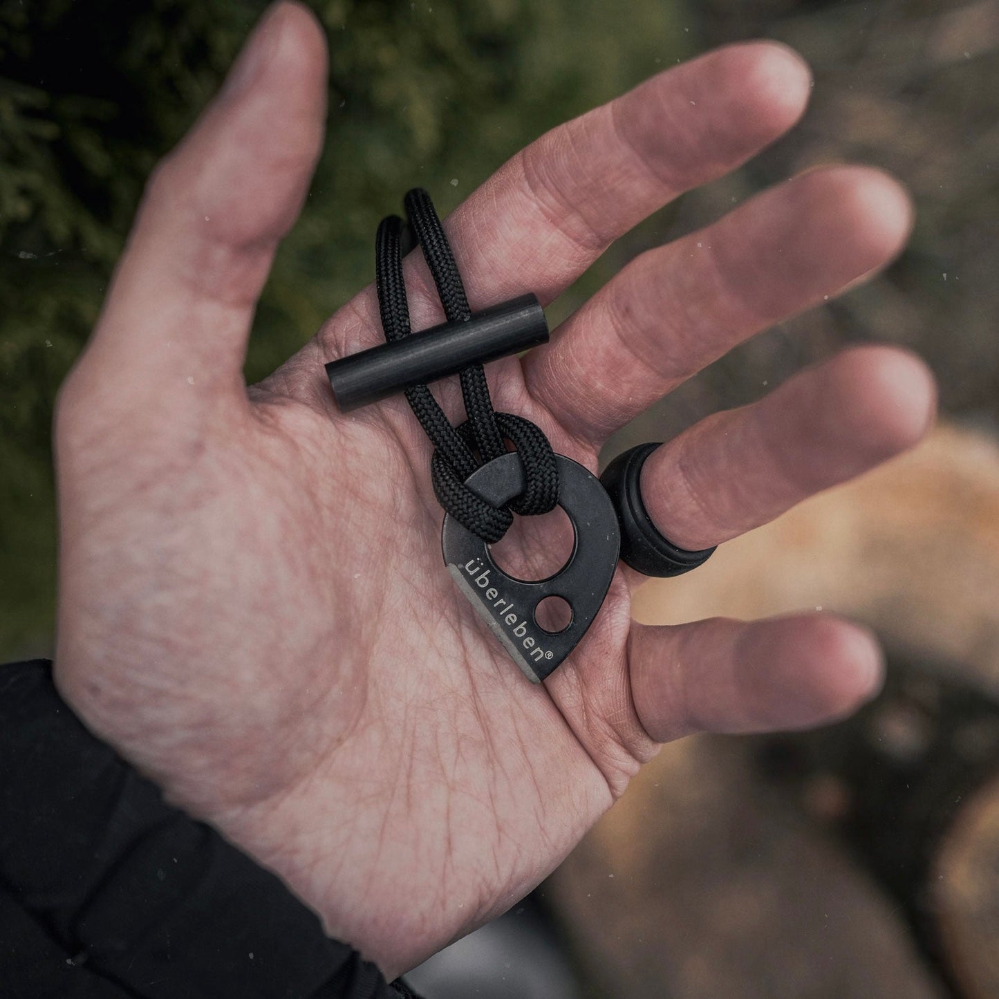 A hand holds the Überleben Leicht Wearable Fire Starter by Überleben, featuring überleben engraved on a metal piece attached to a sturdy paracord, shown against an outdoor backdrop.