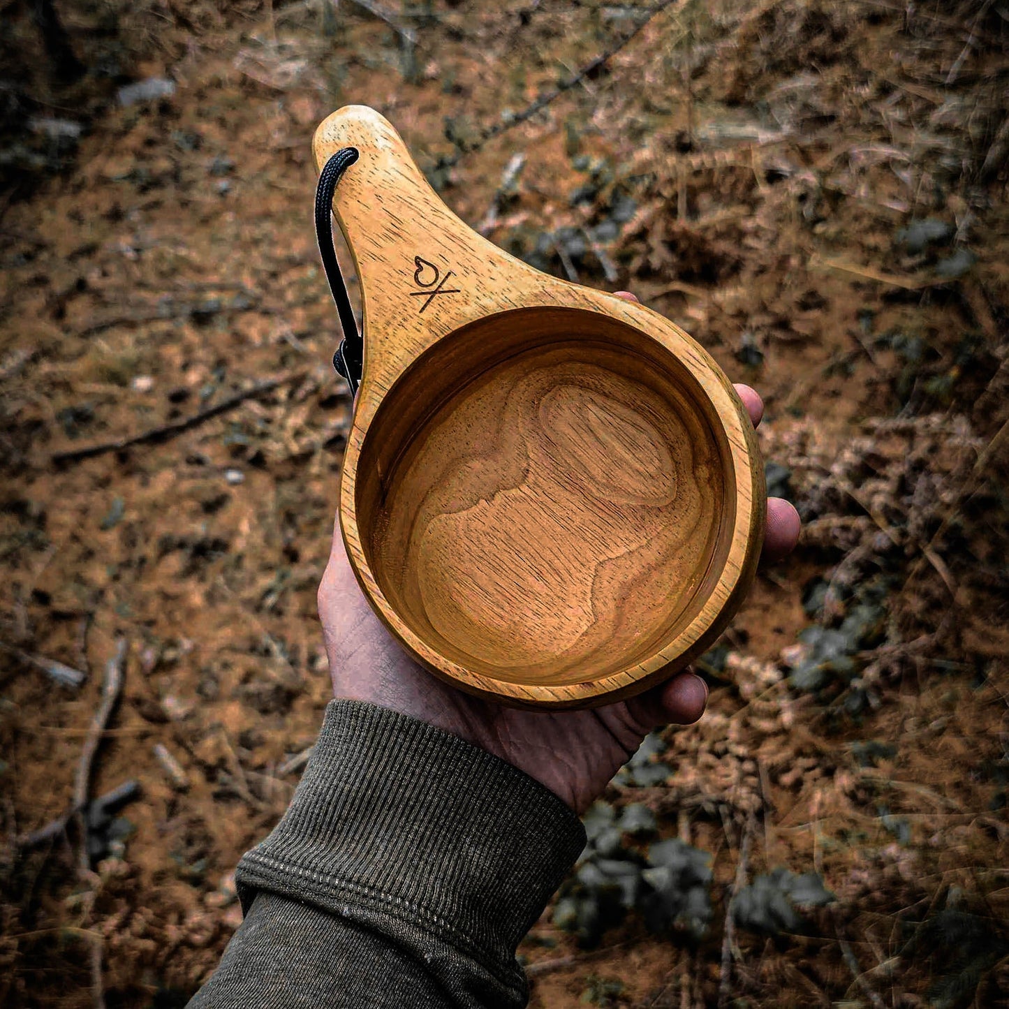 A person in a green sleeve holds Überleben’s The Lore – Oversized Hardwood Kuksa, featuring a handle and leather strap, with an earthy, blurred outdoor background of scattered twigs.