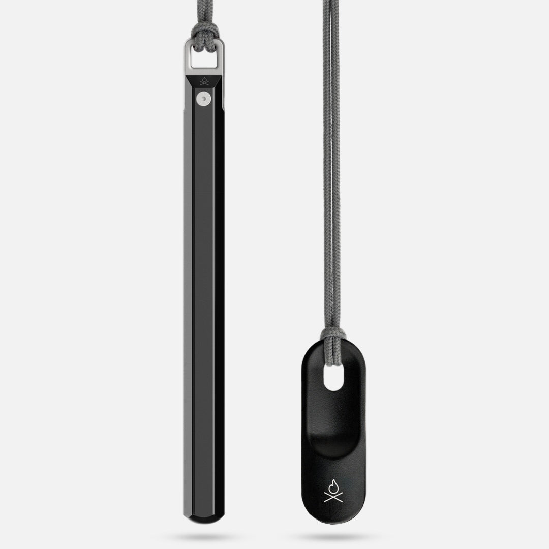 Two sleek, black objects on gray cords hang vertically: Überleben’s Hexå PRO, a slim hex ferro rod fire starter with a button and logo, and an oval pendant featuring a cutout and the same Überleben logo at the bottom.