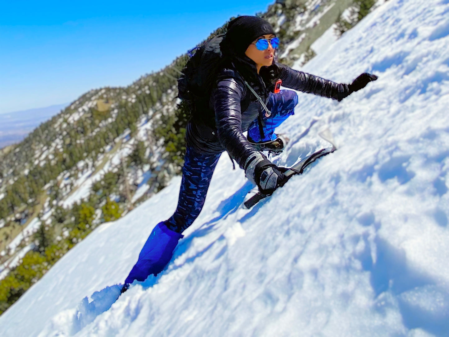 Clad in winter gear and sunglasses, a hiker climbs a snowy mountain with an ice axe, wearing Pike Trail All-Season Waterproof Leg Gaiters—built for the trail, the hunt, and everything in between—amid pine trees under a clear blue sky.