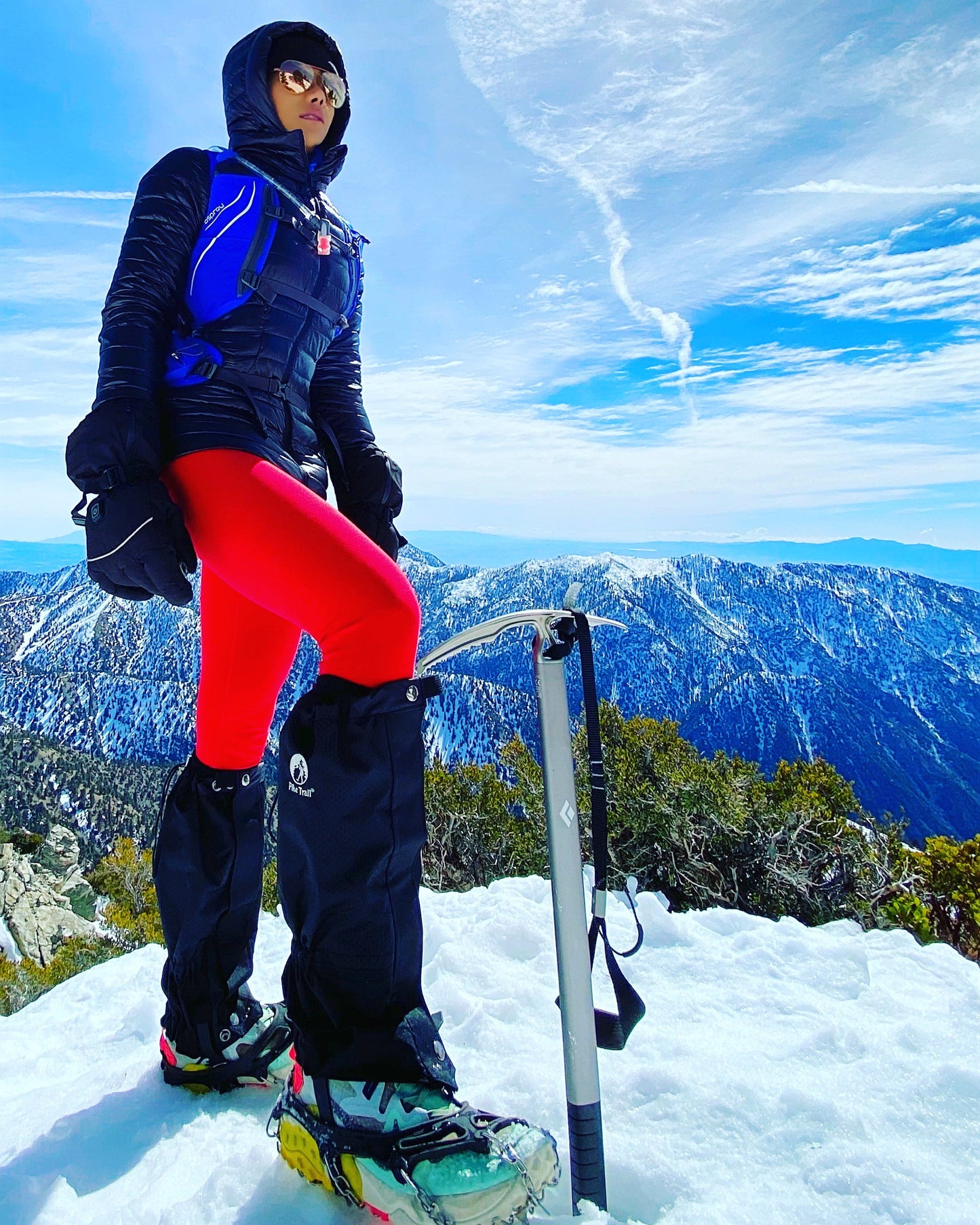 A climber in red pants, black jacket, and snow boots fitted with Pike Trail All-Season Waterproof Leg Gaiters stands atop a snowy peak holding an ice axe, with snowy mountains and a blue sky in the background.