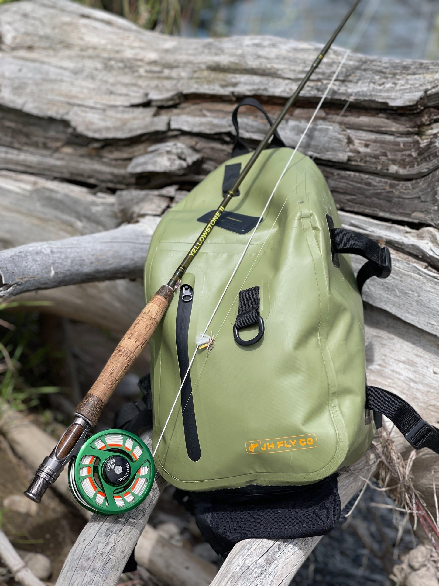 A JHFLYCO 20L Waterproof Sling Pack by Jackson Hole Fly Company, streamlined for the mission, holds a fly rod and reel on weathered driftwood outdoors.
