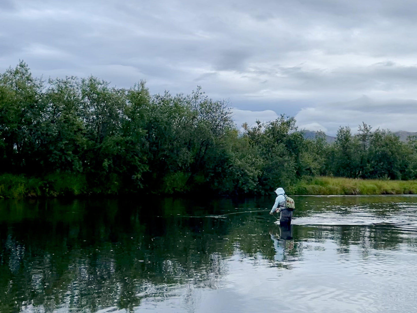 An angler wearing waders and a hooded jacket fly fishes by a tree-lined riverbank under cloudy skies, carrying the Jackson Hole Fly Company JHFLYCO 20L Waterproof Sling Pack—streamlined for the mission—over their shoulder.