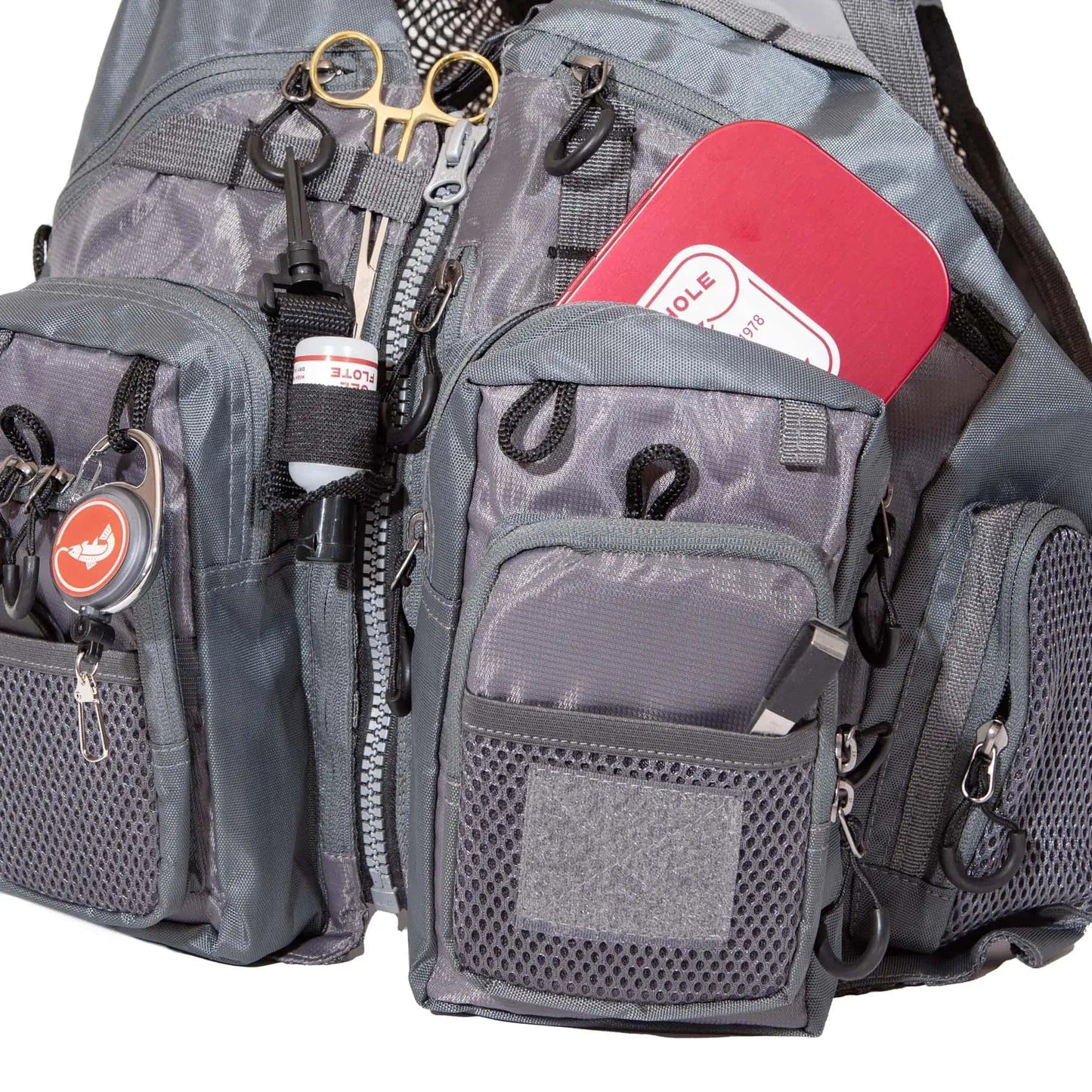 A close-up of the Jackson Hole Fly Company JHFLYCO Adjustable Mesh Fishing Vest in gray, featuring multiple zippered pockets, mesh pouches, and attached gear like scissors, a red and white tin, badge, and black carabiner clip.