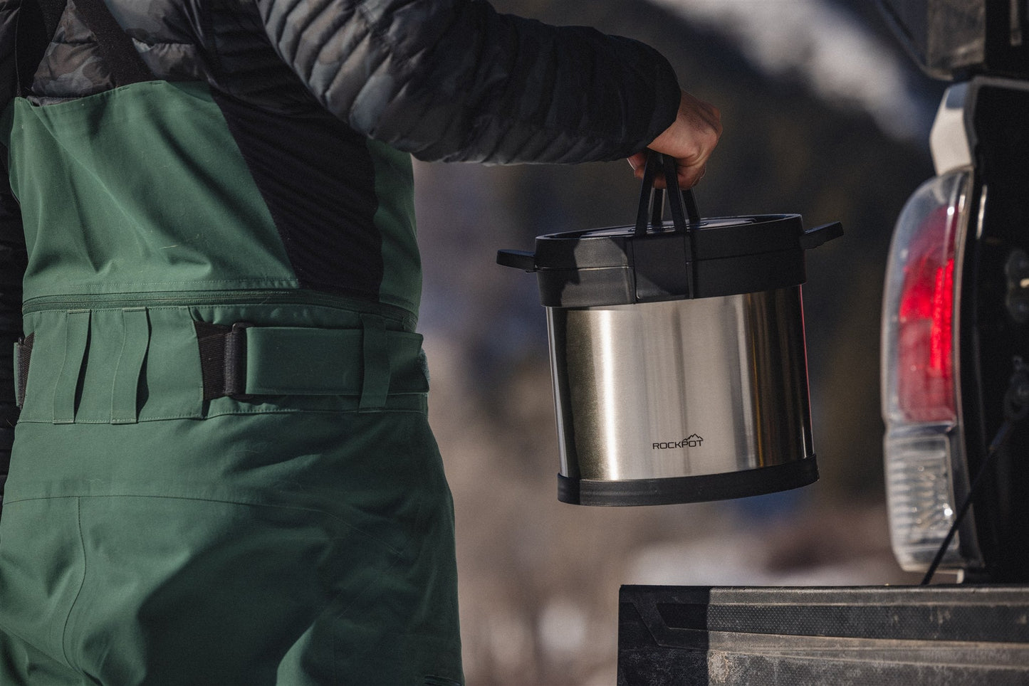 Wearing green overalls and a black jacket, a person lifts the RockPot Mini Bundle – All-In Survival Cooking, No Plug Required by RockPot from a pickup truck outdoors, with mountains and snow in the background.