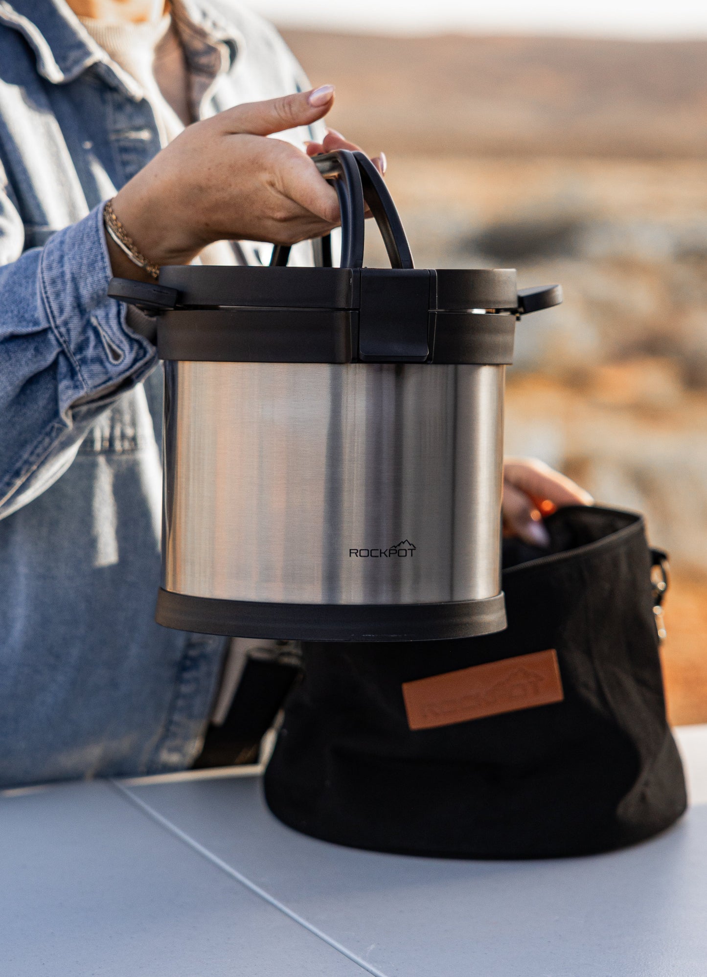 A person in a denim jacket lifts the RockPot Mini Bundle – All-In Survival Cooking, No Plug Required by RockPot from a black bag on an outdoor table, ideal for off-grid cooking with a blurred natural landscape behind.