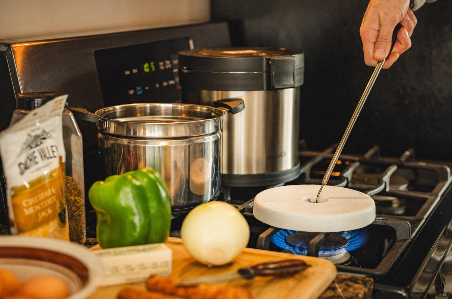 A person cooks on a gas stove with the RockPot Mini Bundle – All-In Survival Cooking, No Plug Required by RockPot, using a metal ladle and white pan. On a wooden board are green bell pepper, onion, eggs, and shredded cheese.