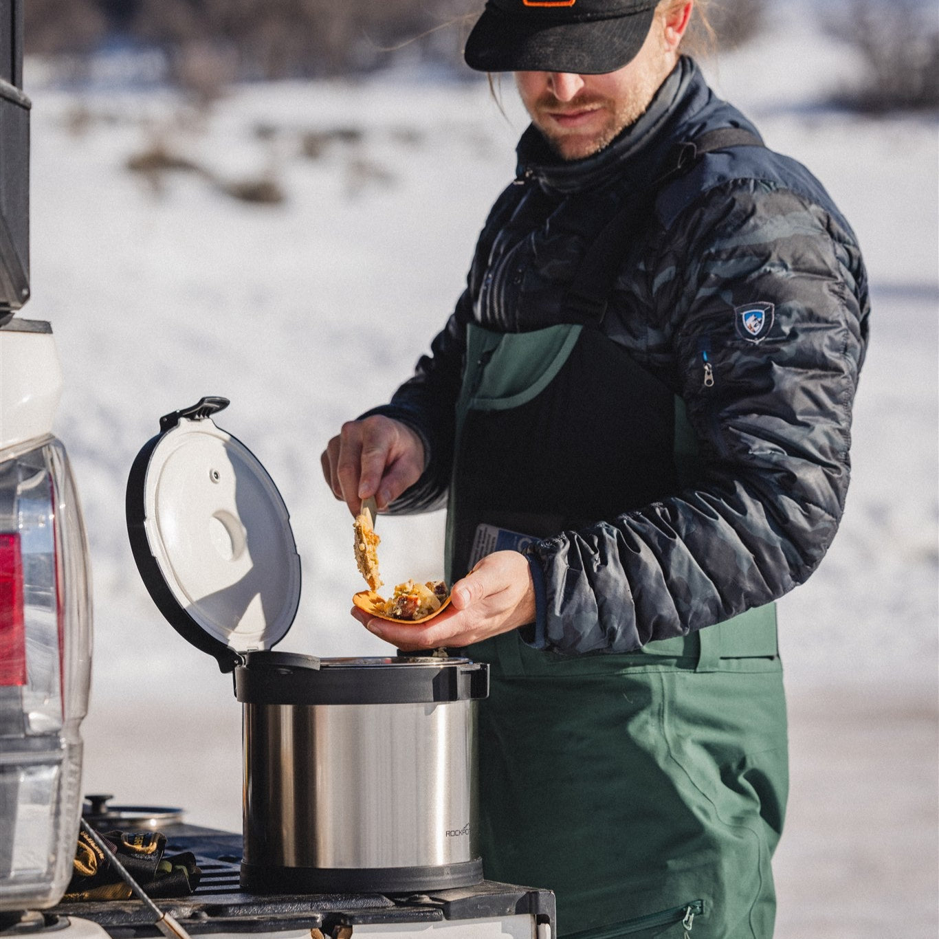 Wearing a green apron and jacket, a person serves food from the RockPot Mini Bundle – All-In Survival Cooking, No Plug Required by RockPot into a bowl beside a vehicle in snowy outdoors.