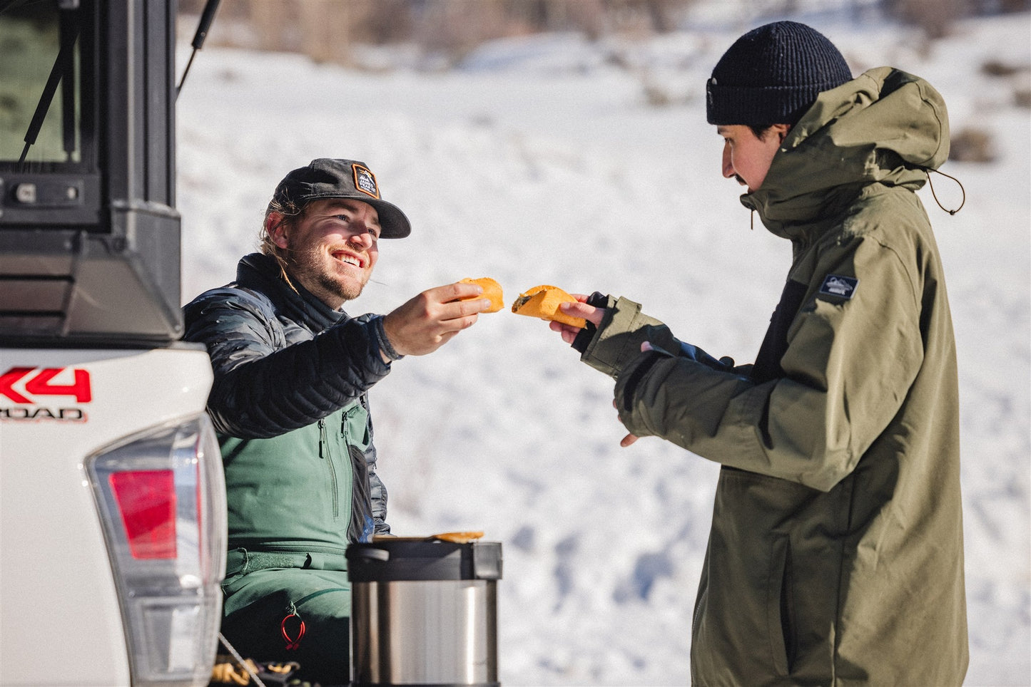 Two people in winter gear stand by an open pickup tailgate in the snow, smiling as they hold grilled cheese sandwiches made with their RockPot Mini Bundle – All-In Survival Cooking, No Plug Required by RockPot, ready to toast them together.
