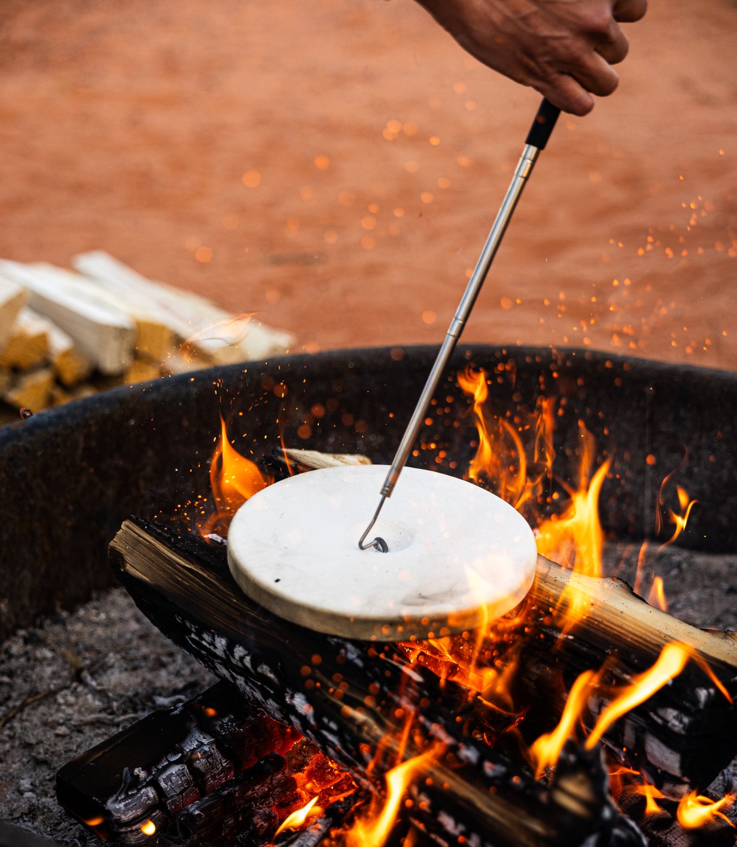 A hand lifts a metal lid from the RockPot Mini Bundle – All-In Survival Cooking, No Plug Required by RockPot, revealing an off-grid cooking system above crackling flames and burning wood, with stacked firewood on the sandy ground.