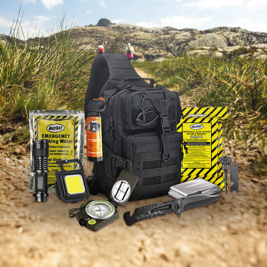 A black Entropy Survival Safe Trail Survive-All Go Bag rests on grass among rocky hills, surrounded by survival essentials—rations, water, flashlight, compass, multi-tool, knife, utility light, and pepper spray—for your emergency kit.
