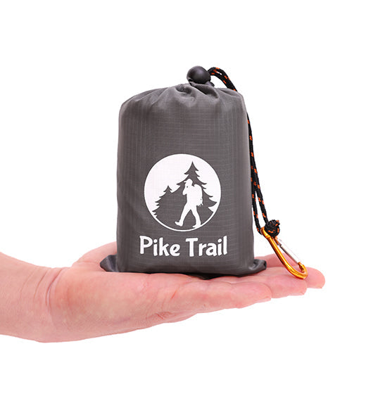 A hand holds the Pike Trail Pocket Blanket’s gray drawstring pouch, branded with a hiker and trees. A yellow carabiner clips on for easy carrying—compact, rugged, and always ready for your next adventure.