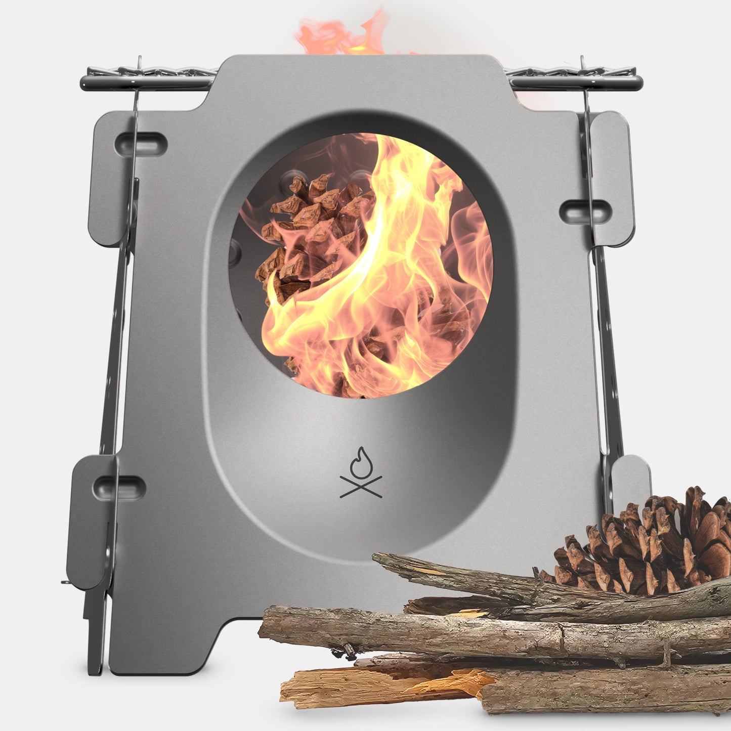 The Überleben Stöker Flatpack Stove by Überleben is a compact metal wood-burning stove with a circular window showing the fire inside. Pinecones and sticks lie beside it on a white background.