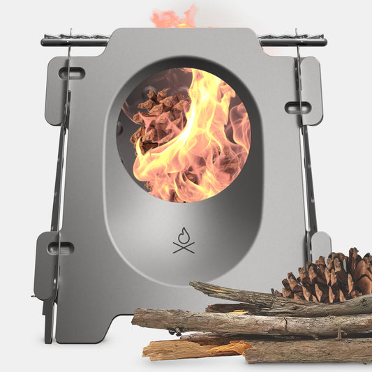The Überleben Stöker Flatpack Stove by Überleben is a compact metal wood-burning stove with a circular window showing the fire inside. Pinecones and sticks lie beside it on a white background.