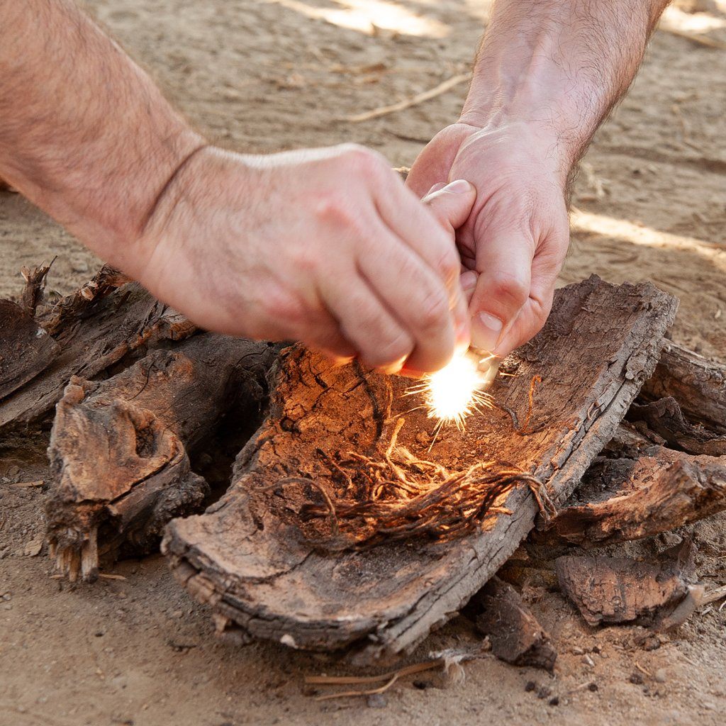 Two hands use a fire starter to ignite dry twigs, relying on TITAN Survival’s SurvivorCord® Boot Laces—incorporating Kevlar, fishing line, and a firestarter—for added security in an outdoor emergency.