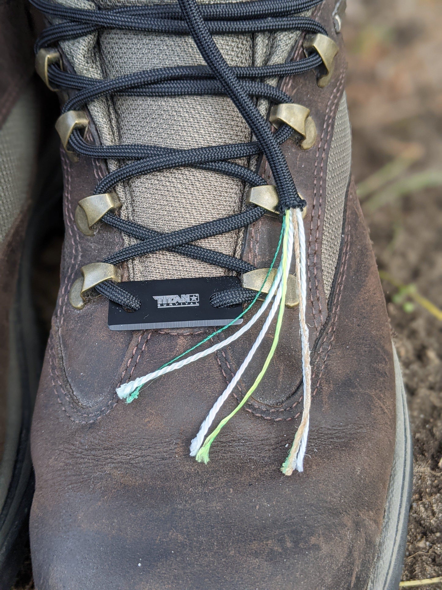Close-up of a worn brown hiking boot with TITAN Survival’s SurvivorCord® Boot Laces – Firestarter, Kevlar, Fishing Line, threaded through metallic eyelets. White, green, and tan inner strands are visible under the laces near the toe.