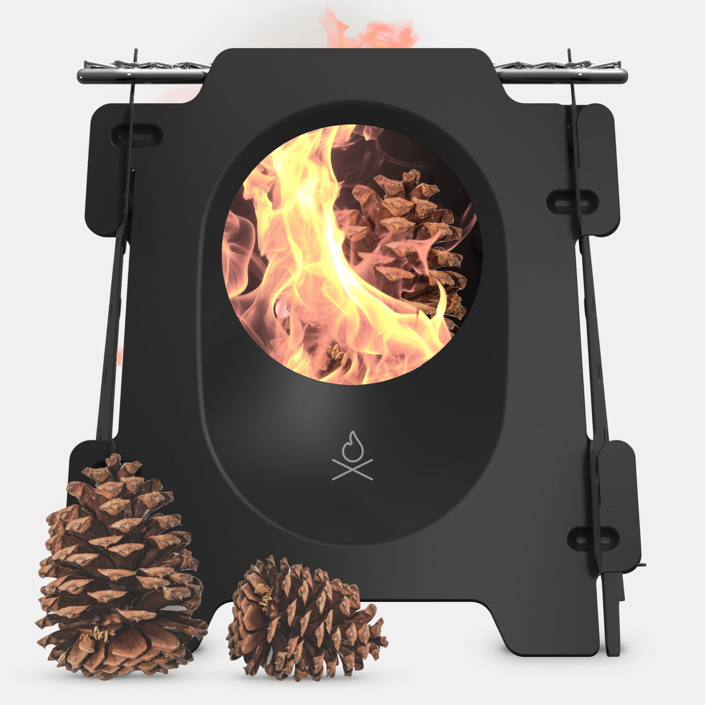The Überleben Stöker Flatpack Stove by Überleben is a black rectangular wood-burning camp stove with a circular window showing flames and burning pine cones inside, while two more pine cones sit in front on a white background.