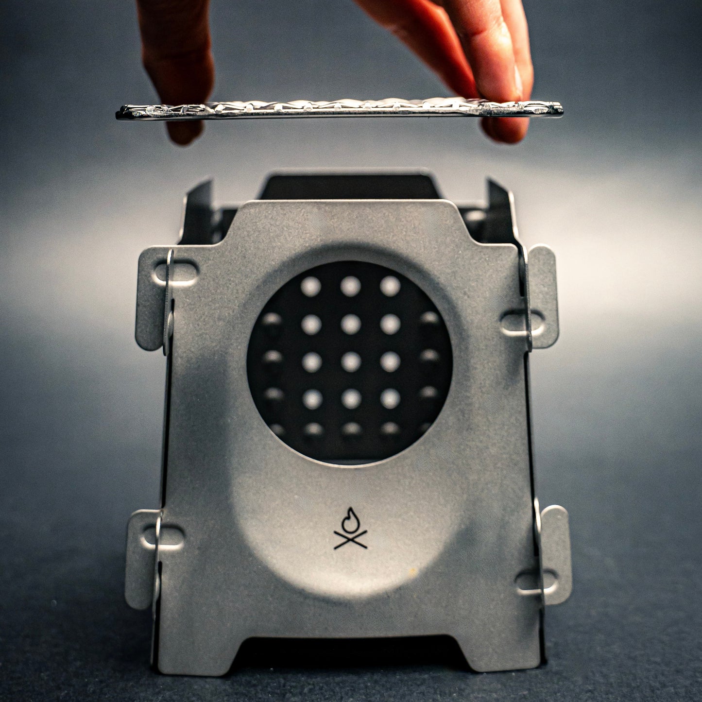 A hand lifts the lid off the Überleben Stöker Flatpack Stove, a compact metal wood-burning stove with a circular vent and flame logo from Überleben, showcasing this survival essential against a dark background.