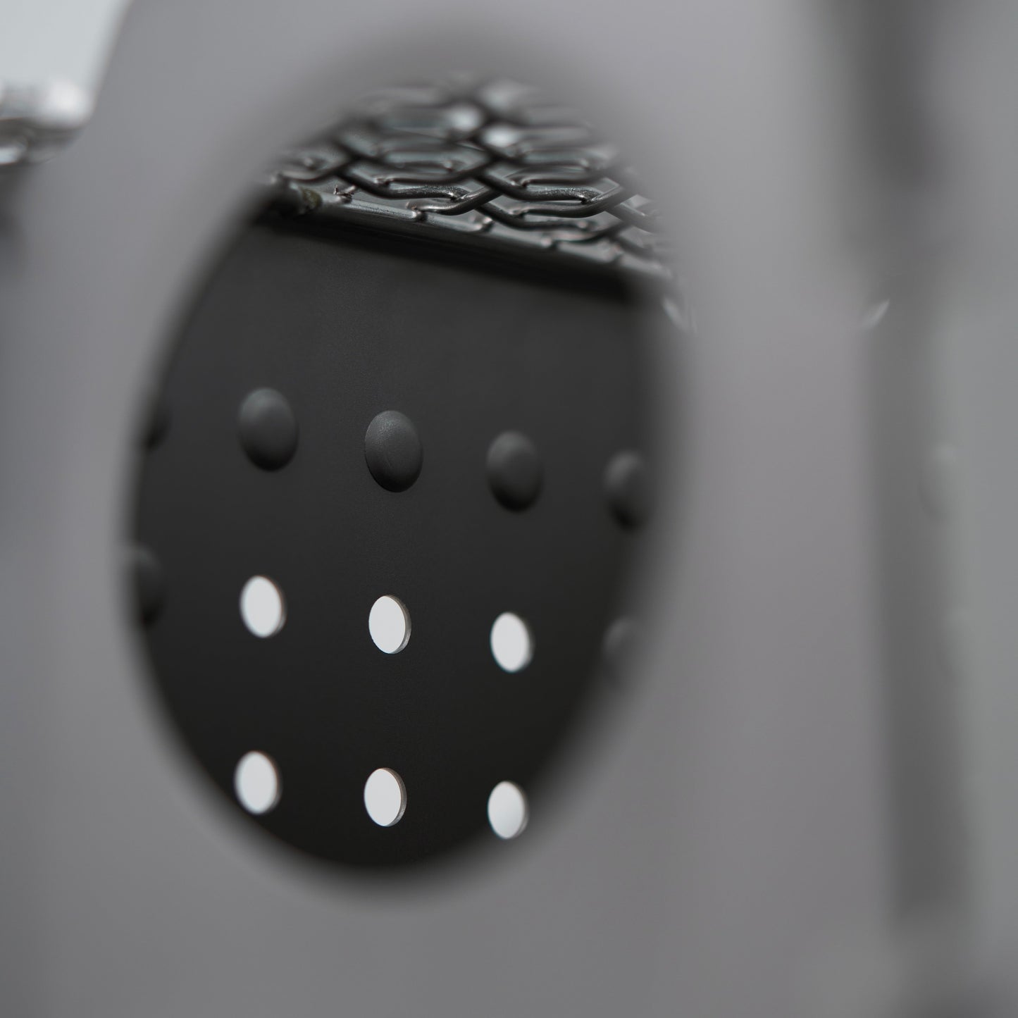 A close-up of the Überleben Stöker Flatpack Stove by Überleben shows its circular metal surface with evenly spaced holes, partially obscured in the foreground. The out-of-focus background displays a subtle grid-like pattern.