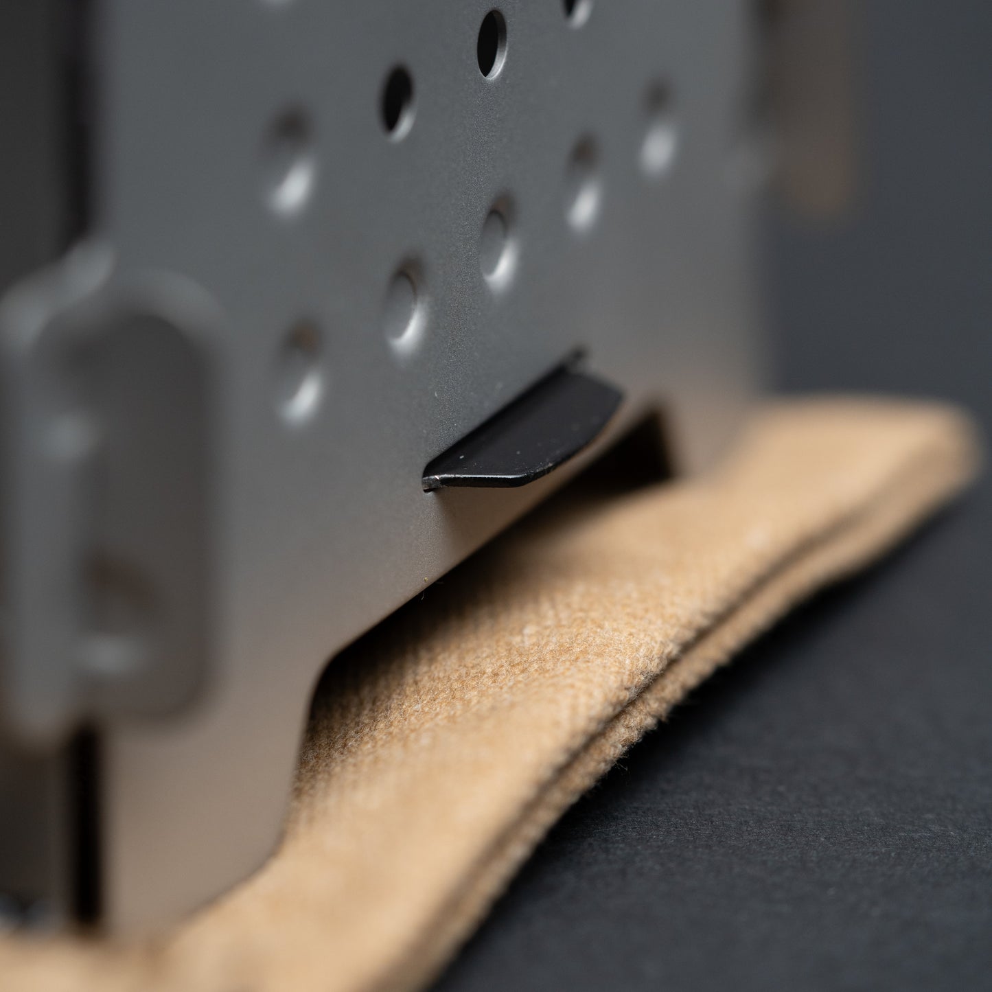 A close-up of the Überleben Stöker Flatpack Stove by Überleben, a metallic survival tool with round holes and a black tab, resting on beige fabric against a dark background.