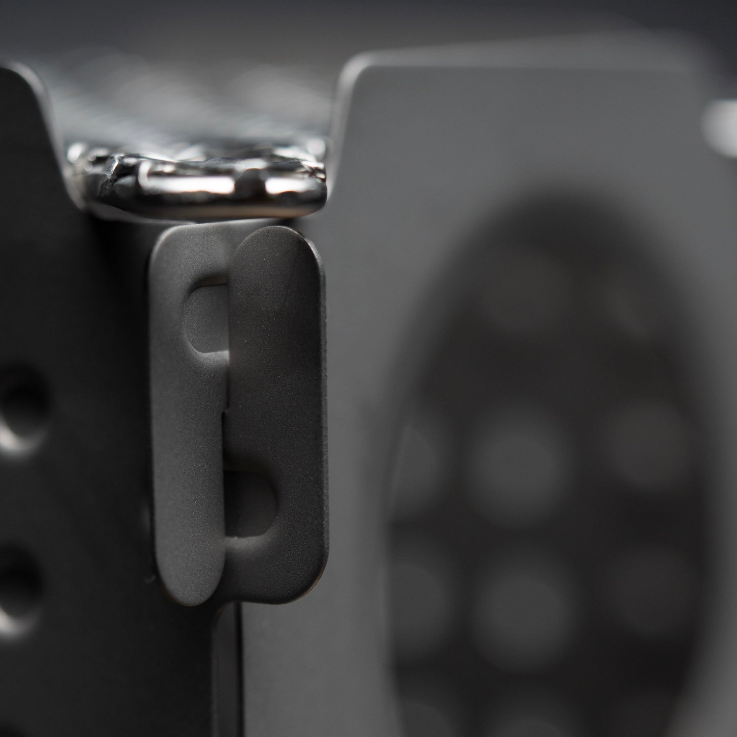 Close-up of a matte black metal latch on the Überleben Stöker Flatpack Stove by Überleben, shown attached to its perforated surface with a blurred patterned background.
