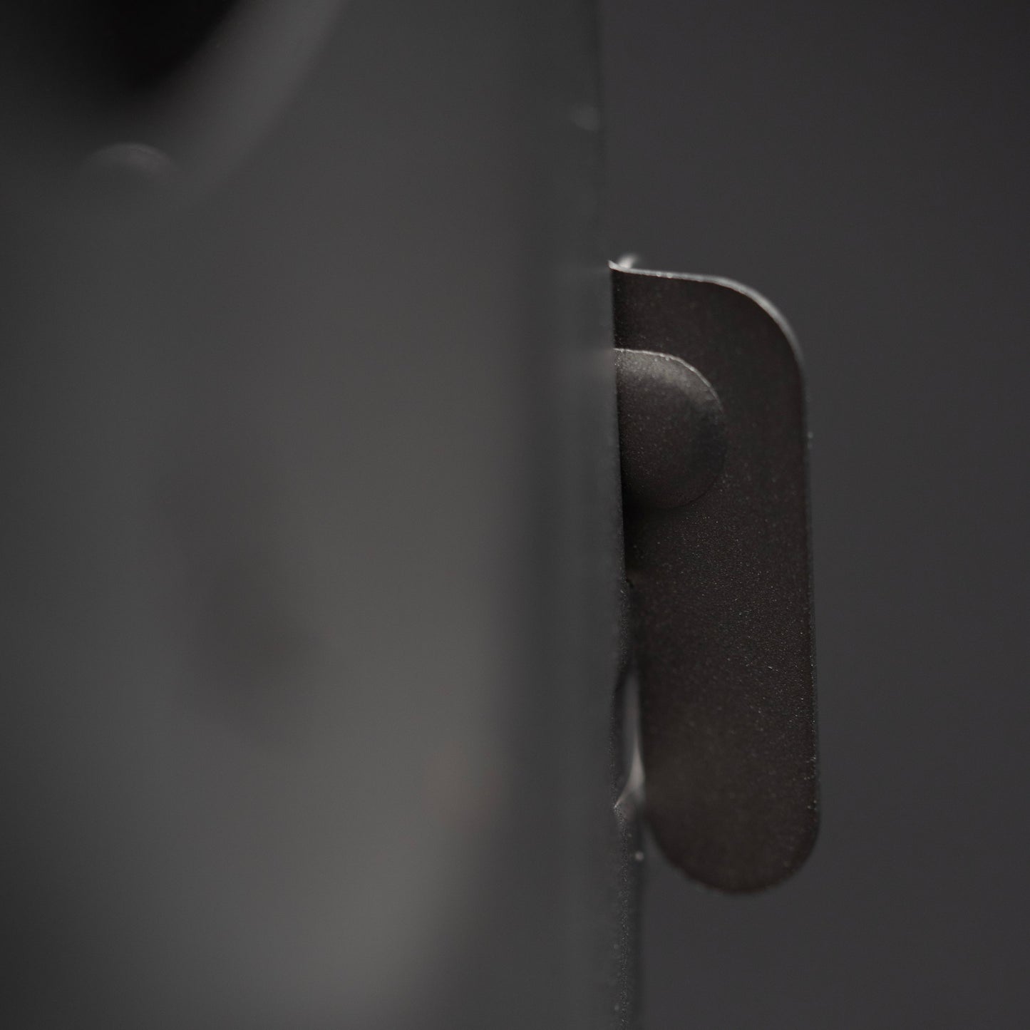 Close-up of a dark metal latch protruding from the smooth surface of the Überleben Stöker Flatpack Stove by Überleben, with a blurred, dark background.