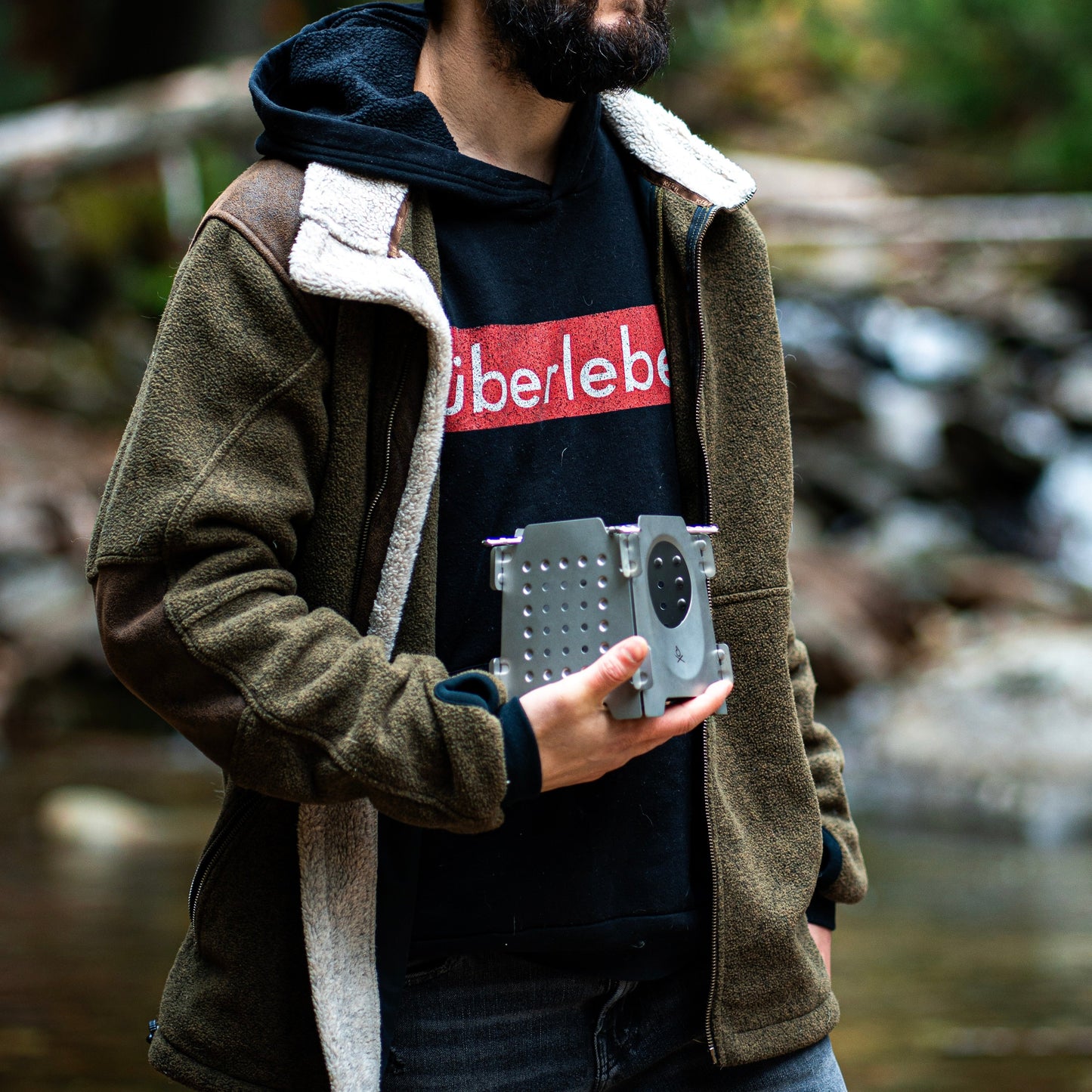 Outdoors near a river with a blurred forest behind, someone in a brown jacket and black Überleben hoodie holds a gray drone controller, ready to explore or try their Überleben Stöker Flatpack Stove. Their face is partially out of frame.