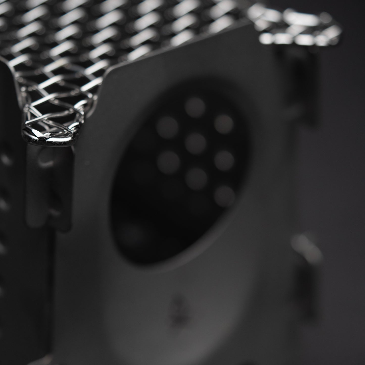 Close-up of the Überleben Stöker Flatpack Stove by Überleben, featuring a metallic, perforated grid top and side opening. The image highlights this survival tools textured finish and geometric design against a dark, blurred background.