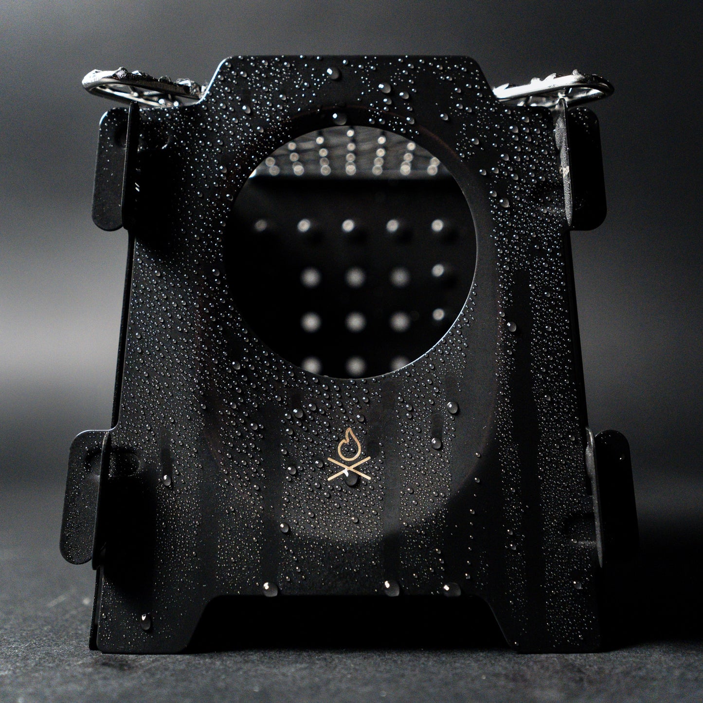 The Überleben Stöker Flatpack Stove by Überleben—a black metal stove with a circular cutout, water droplets on its surface, and a small campfire logo at the bottom—sits against a dark background.