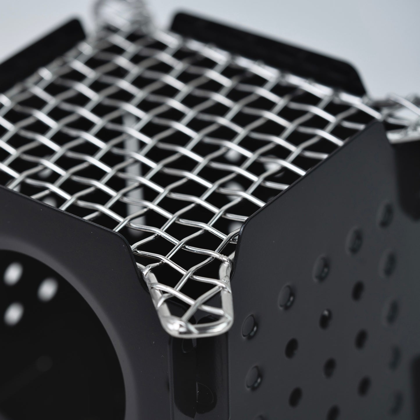 A close-up of the Überleben Stöker Flatpack Stove by Überleben, highlighting its black metal body with a perforated side and silver wire mesh top for detailed construction.