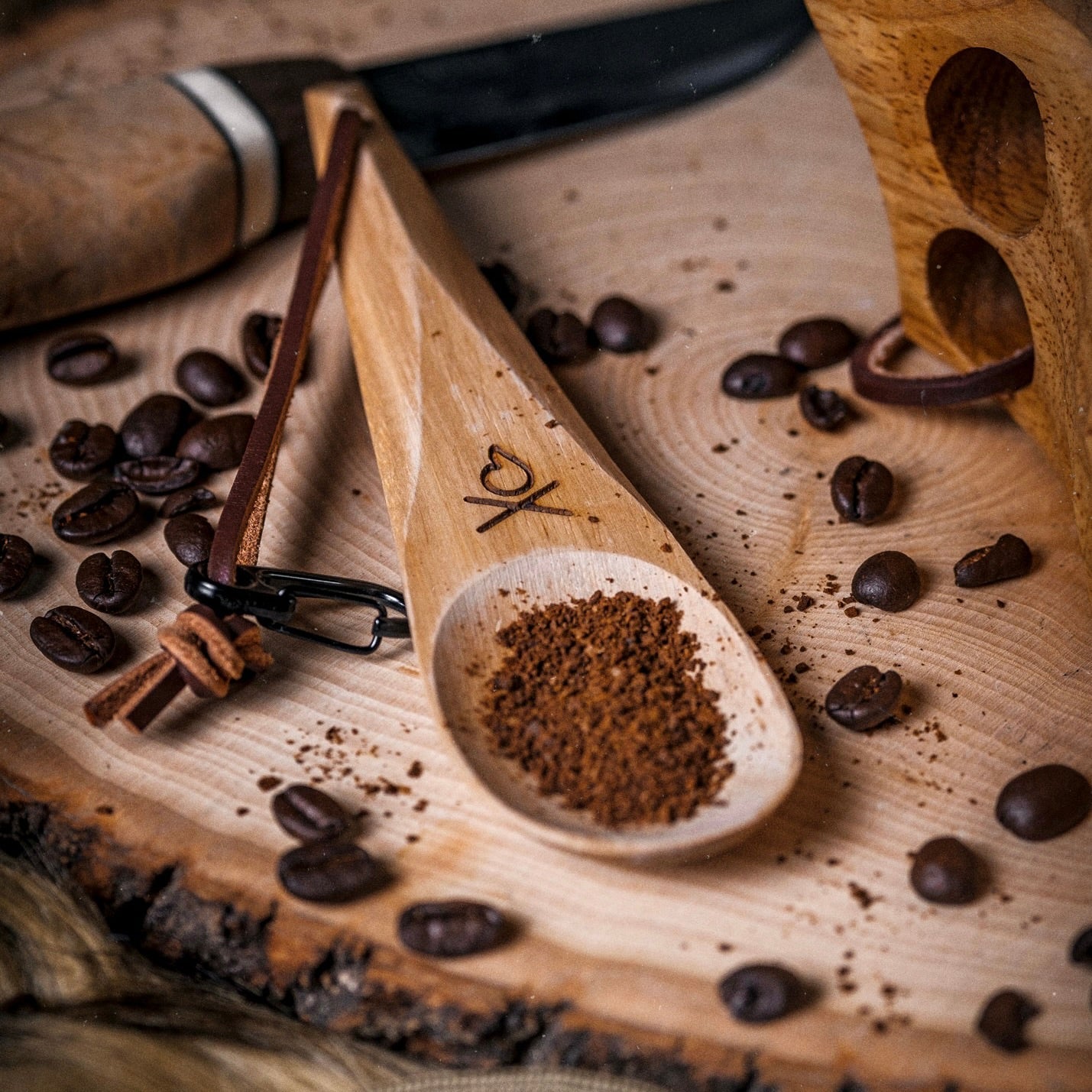 The Überleben Kanu – Hardwood Camping Spoon by Überleben rests on a tree slice with ground coffee and a heart design, surrounded by coffee beans. A wooden-handled knife and part of a wooden holder are also visible.