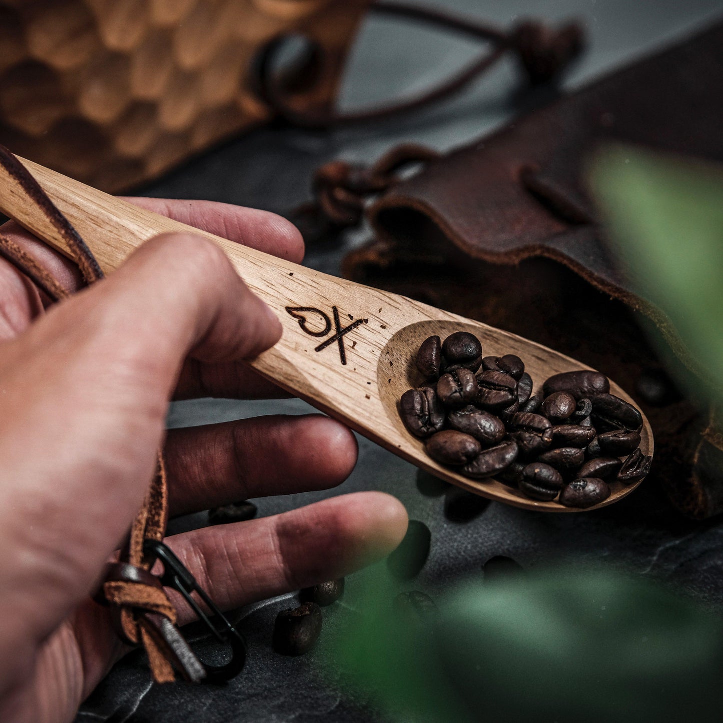 A hand holds the Überleben Kanu – Hardwood Camping Spoon by Überleben, filled with dark roasted coffee beans. A leather pouch and green leaves sit in the background, adding a rustic and cozy touch.