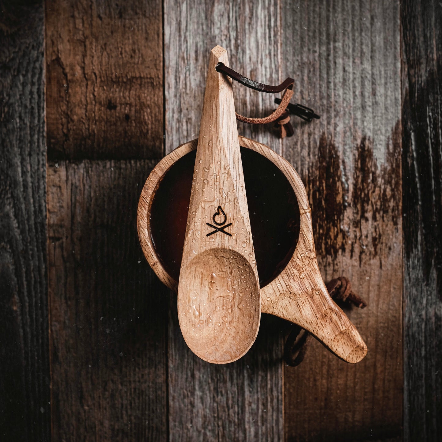 An Überleben Kanu Hardwood Camping Spoon rests across a wooden cup filled with dark liquid on a rustic wood surface.