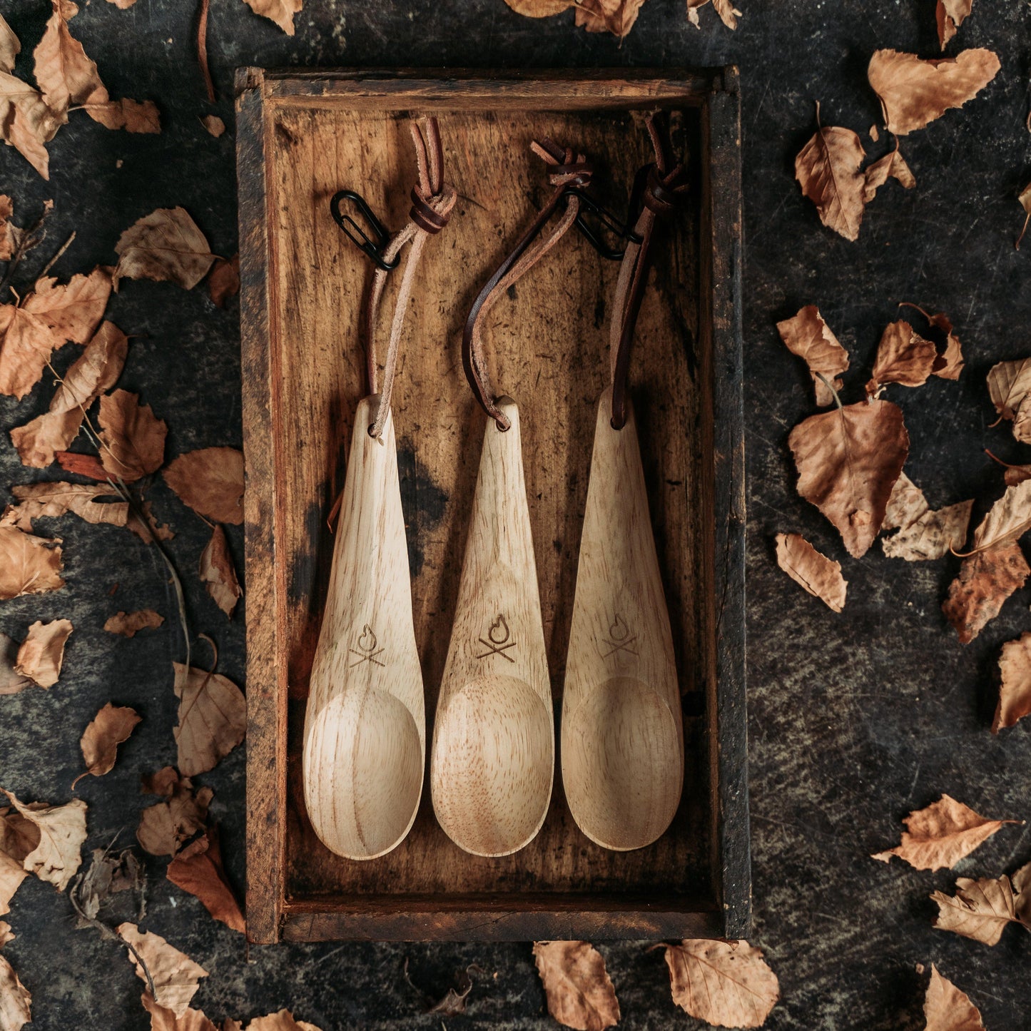 Three Überleben Kanu Hardwood Camping Spoons by Überleben, each with a leather strap, are arranged in a rustic wooden tray and surrounded by dry brown leaves on a dark background.
