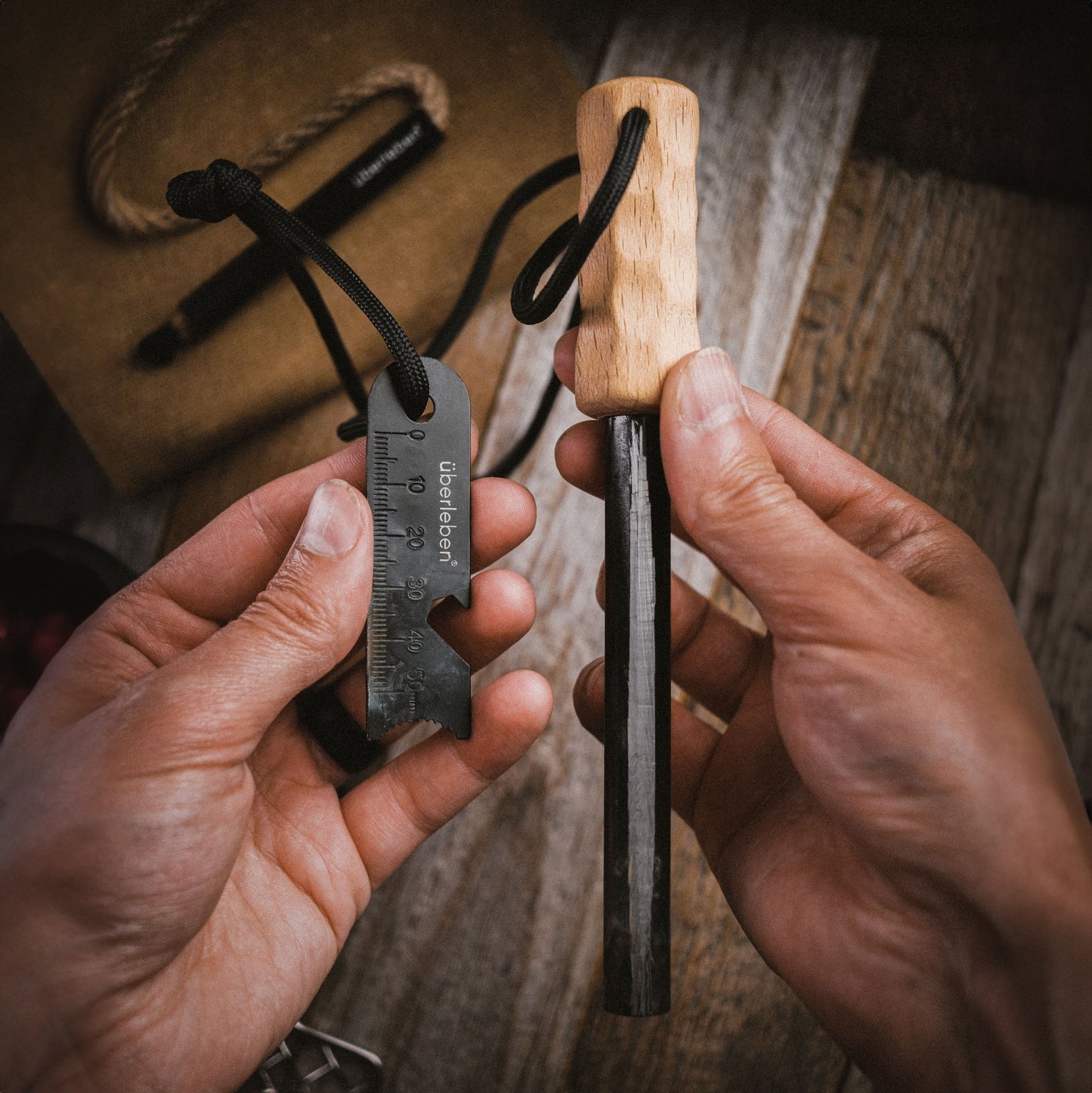 Two hands hold the Überleben Kräftig Grip (PRO) Heavy-Duty Ferro Beast fire starter with wooden handle and multi-tool striker featuring measurement marks by Überleben, ready for use over a rustic wooden table.